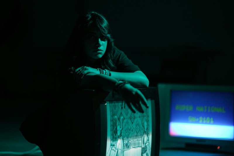 A young woman leaning on the top of a cathode-ray television in a dark room, a second set beside her showing a bright blue screen.