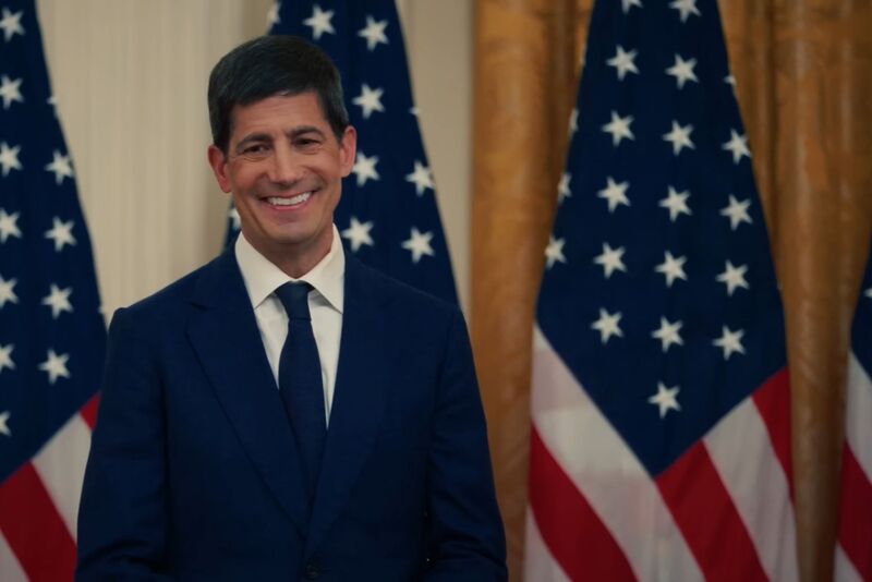 Kevin Warsh, smiling, in a dark suit and tie, stands in front of a row of American flags and gold curtains.