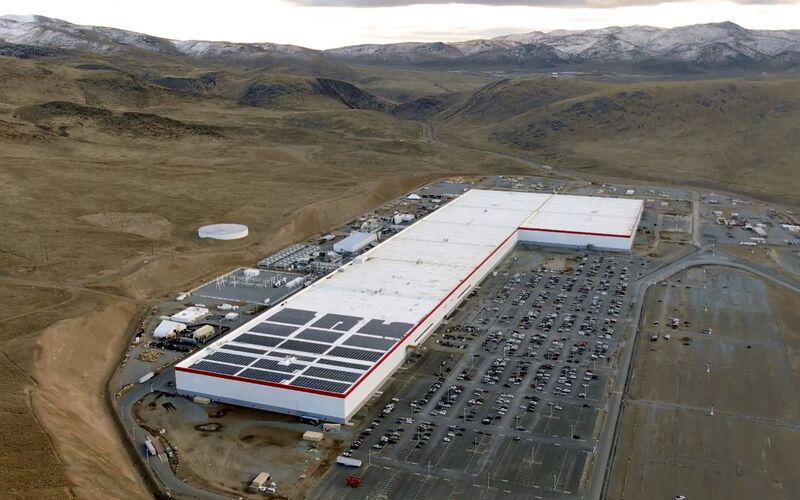 An aerial view of a long white factory building with a red stripe and solar panels on its roof, beside a large car park, in a desert valley with snow-capped hills behind.