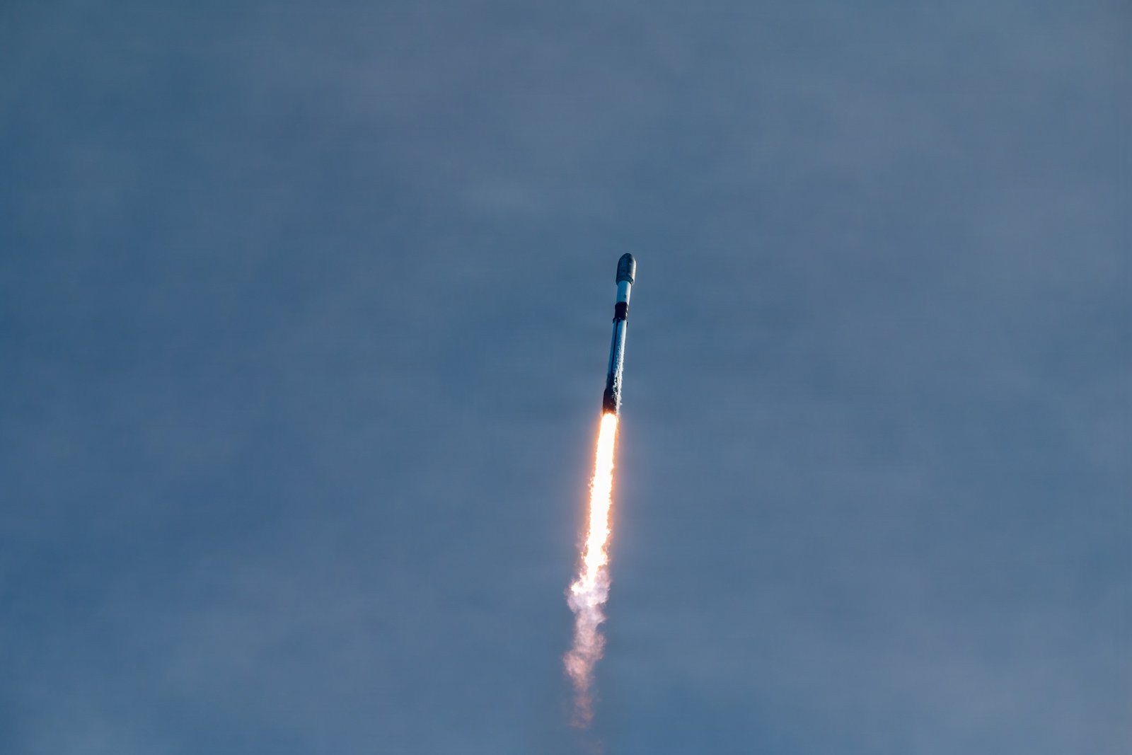 A white Falcon 9 rocket climbs through a pale blue sky on a bright column of orange flame trailing grey exhaust.