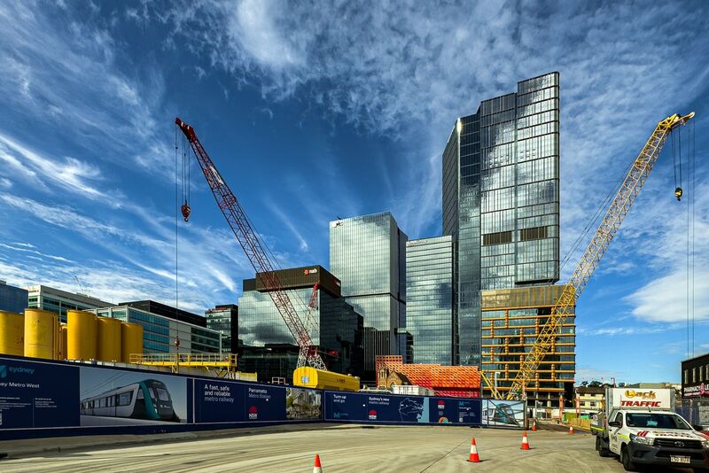 Two construction cranes stand beside glass office towers and a building still in its steel frame, behind blue site hoardings for the Sydney Metro West project, under a sky streaked with cloud.
