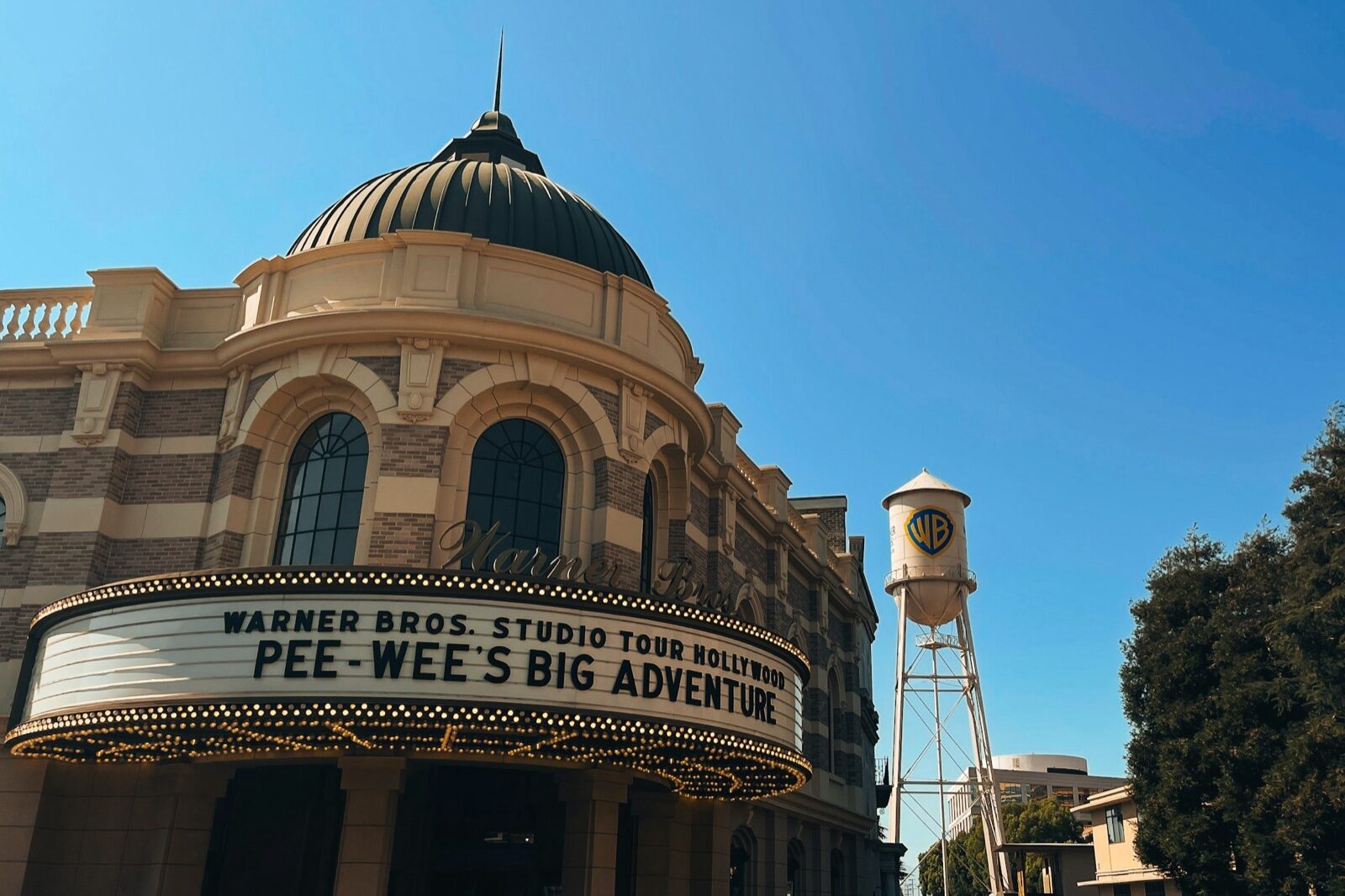 A domed building with a lit marquee reading Warner Bros. Studio Tour Hollywood at the studio lot in Burbank, with the Warner Bros. water tower behind it under a clear blue sky.