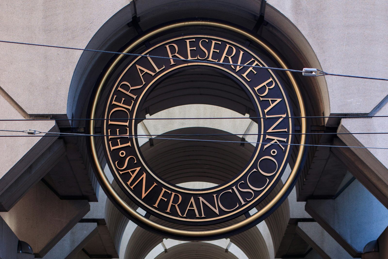 A large bronze ring lettered Federal Reserve Bank San Francisco hangs at the entrance of a vaulted concrete arcade, with overhead cables crossing in front of it.