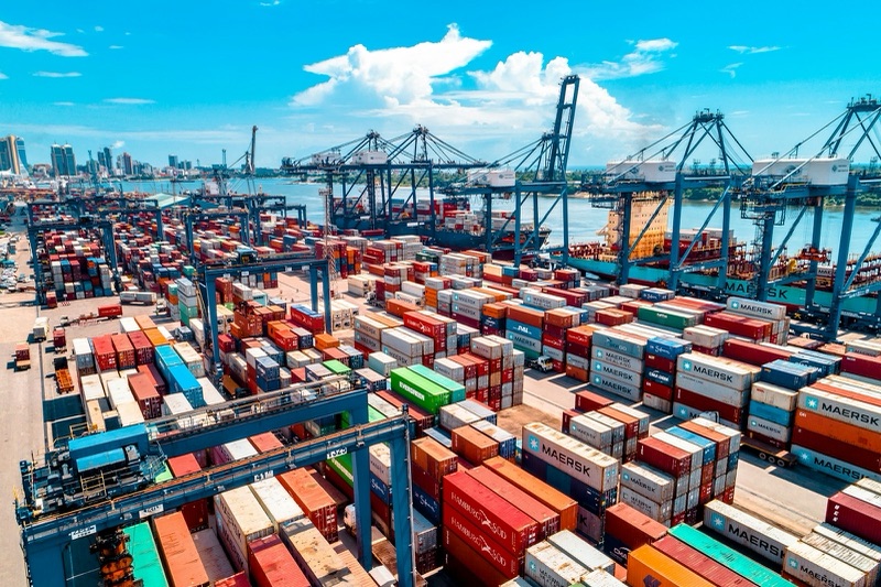 Rows of stacked shipping containers in a port yard, with tall blue cranes along the quayside against a bright blue sky.