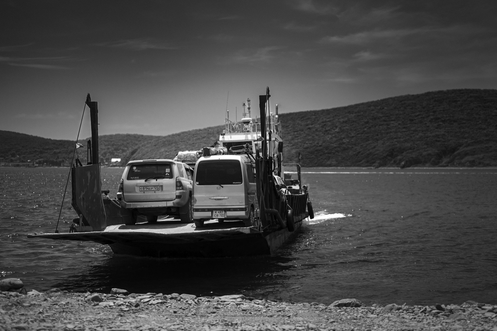 Two vehicles on the open deck of a small ferry crossing a bay, in black and white.