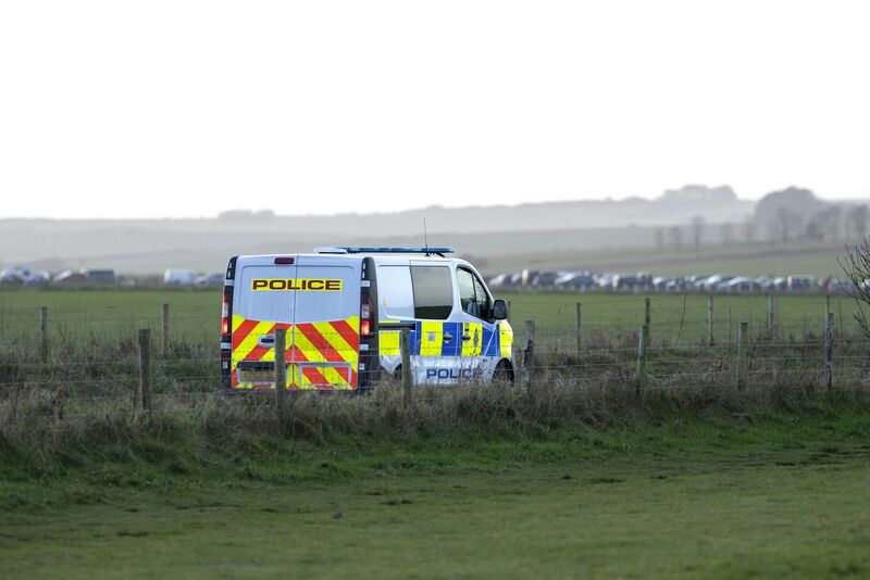 A British police van with yellow and blue reflective markings parked in a green field behind a wire fence, with flat farmland and a line of parked vehicles on the horizon under a pale sky.