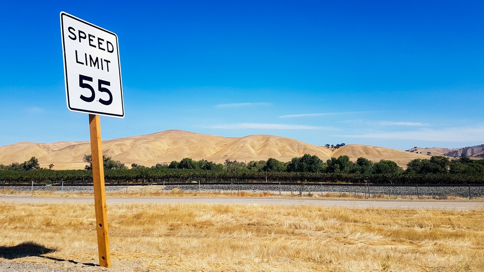A Speed Limit 55 sign beside a road, dry hills behind it under a clear sky.