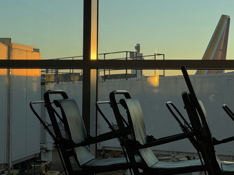 Empty seats at an airport departure gate in silhouette against a floor-to-ceiling window at dusk.