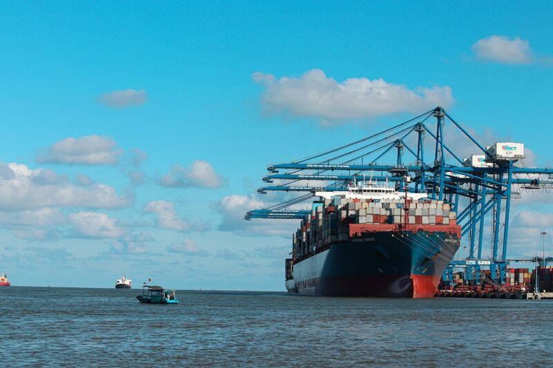 A loaded container ship berthed under blue ship-to-shore cranes at a container terminal in Hai Phong, Vietnam, with a small fishing boat in the foreground.