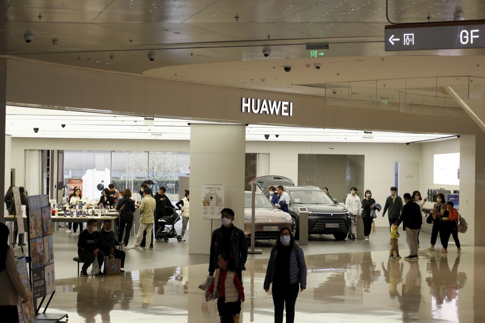 Shoppers, several wearing face masks, walk past a large Huawei store with cars on display inside a bright shopping centre with Chinese signage.