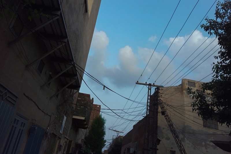 Overhead electricity wires and a utility pole strung between brick buildings on a narrow street in Lahore, under a blue sky with white clouds.