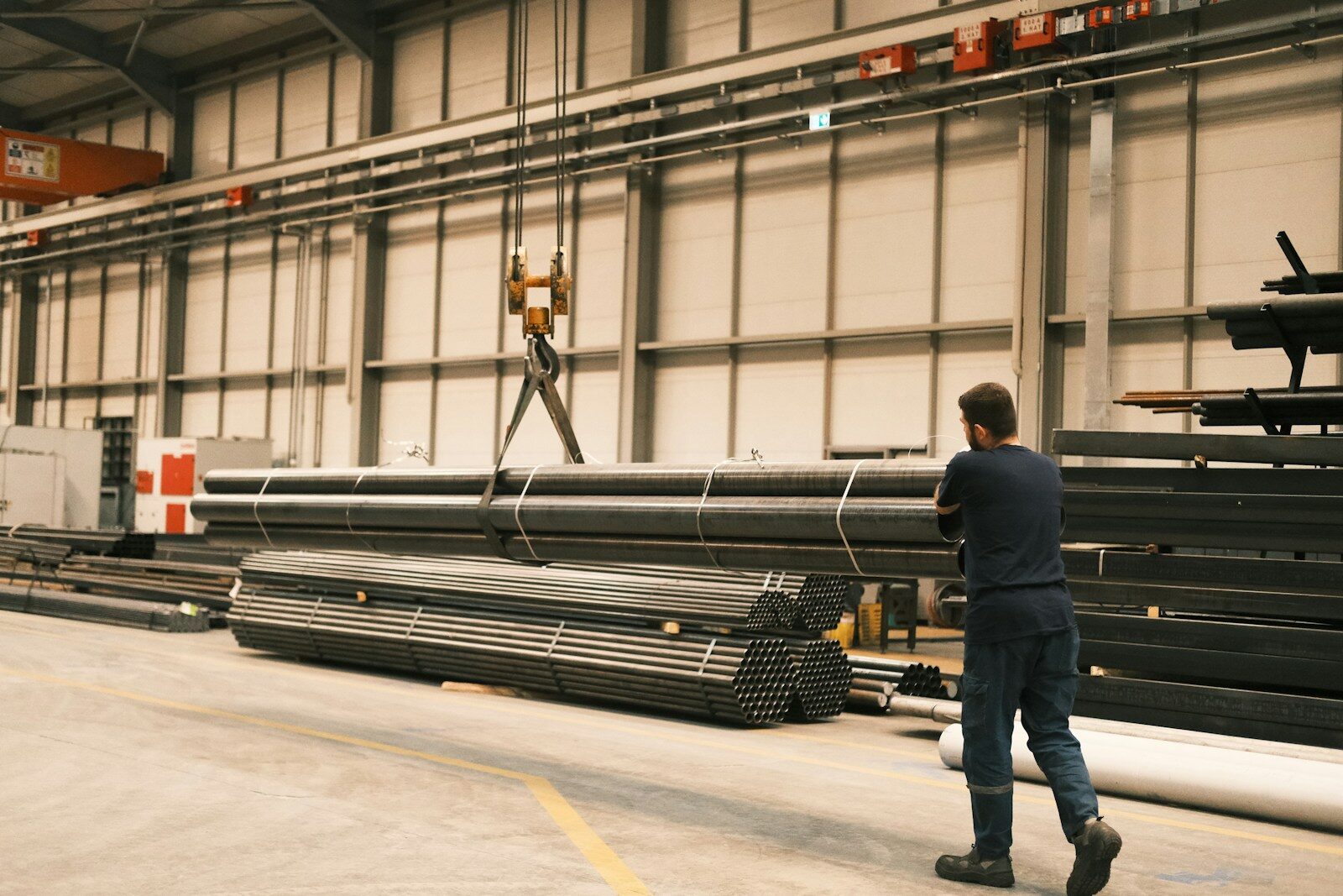 A worker guiding a crane lifting a bundle of steel tube in a distribution warehouse.