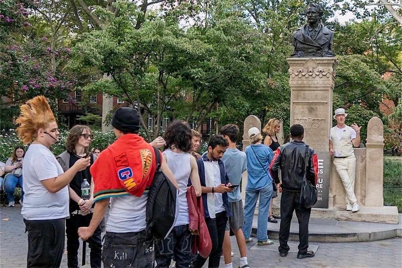 A group of young people stands on the paving of Washington Square Park, some talking and some looking at phones, while a man in a white hat, white waistcoat and white trousers leans against the stone base of a monument topped by a bronze bust and speaks to them. Trees and brick townhouses are behind.