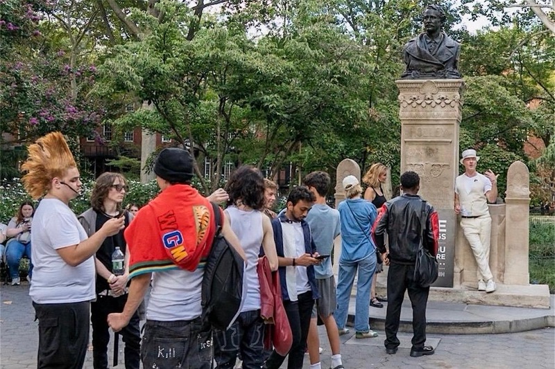 A group of young people stands on the paving of Washington Square Park, some talking and some looking at phones, while a man in a white hat, white waistcoat and white trousers leans against the stone base of a monument topped by a bronze bust and speaks to them. Trees and brick townhouses are behind.