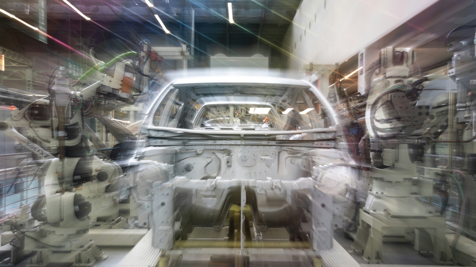 A long-exposure view of robotic arms around a car body on an automated assembly line.