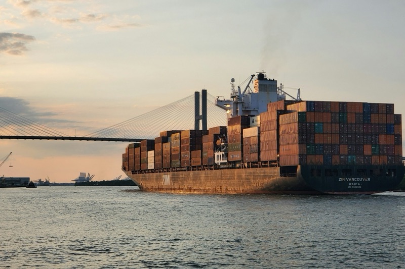 A container ship stacked high with shipping containers sails on the Savannah River at sunset, with a cable-stayed bridge and port cranes in the background.