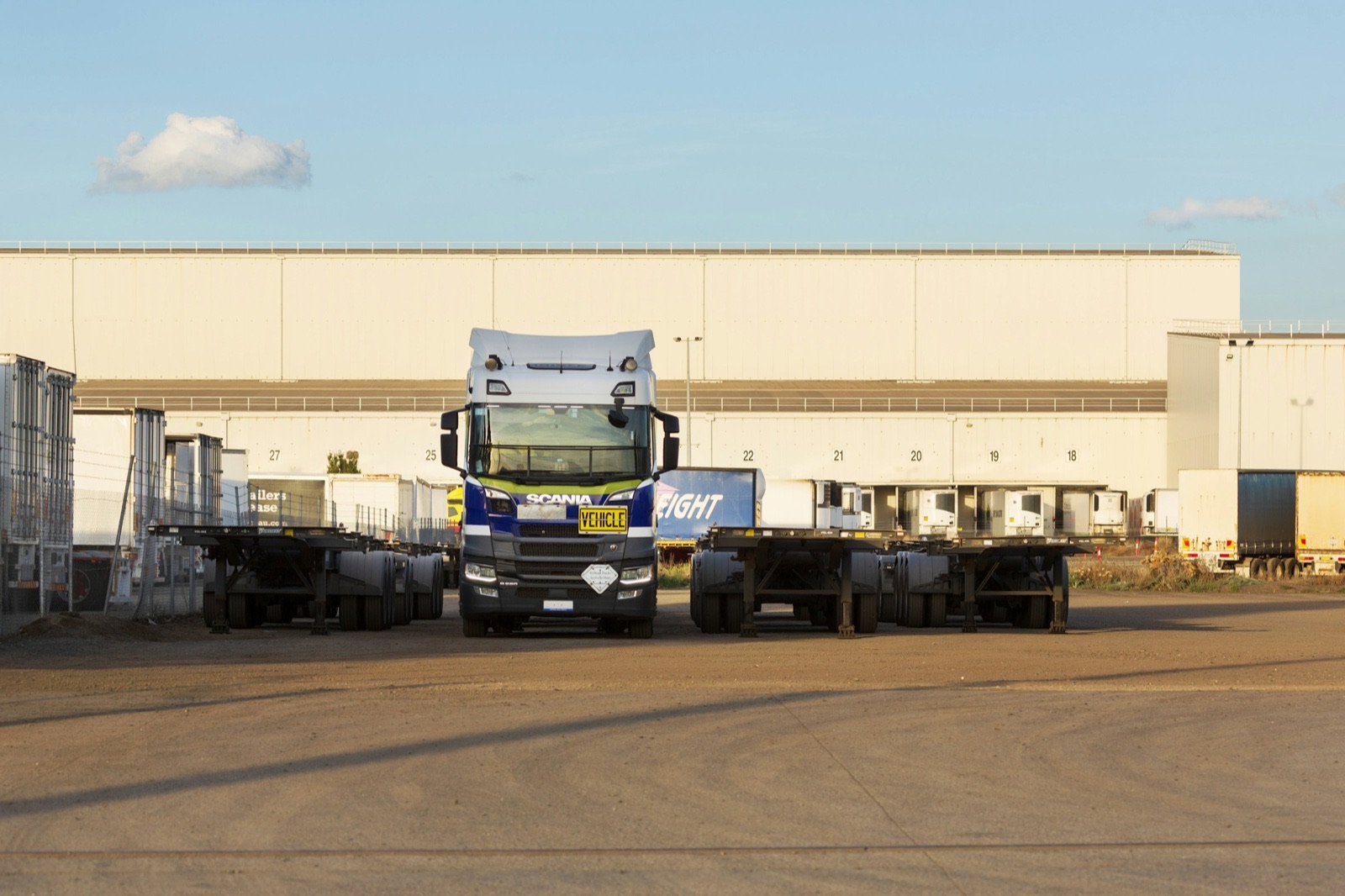 A white truck cab stands between two empty flatbed trailers in a gravel yard in front of a long warehouse with loading bays, under a blue evening sky.