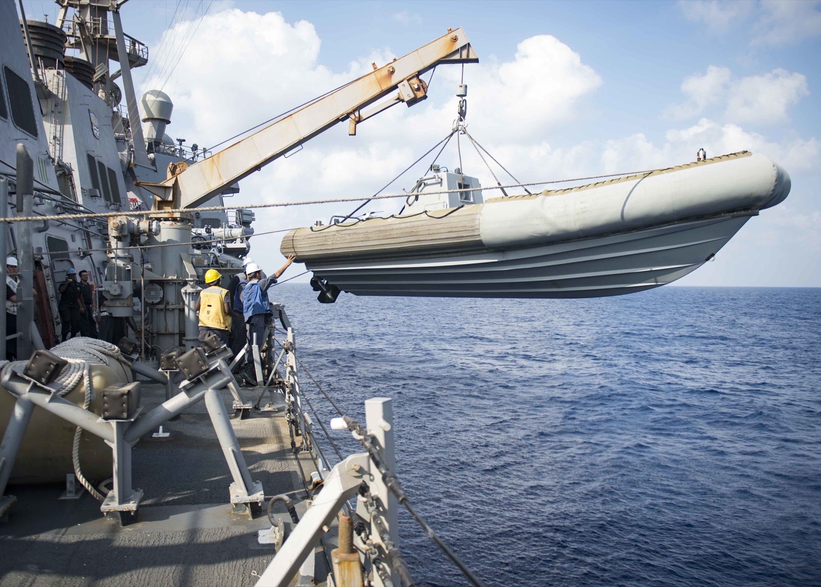 Sailors on the deck of a US Navy destroyer lower a rigid-hull inflatable boat from a davit over open water.