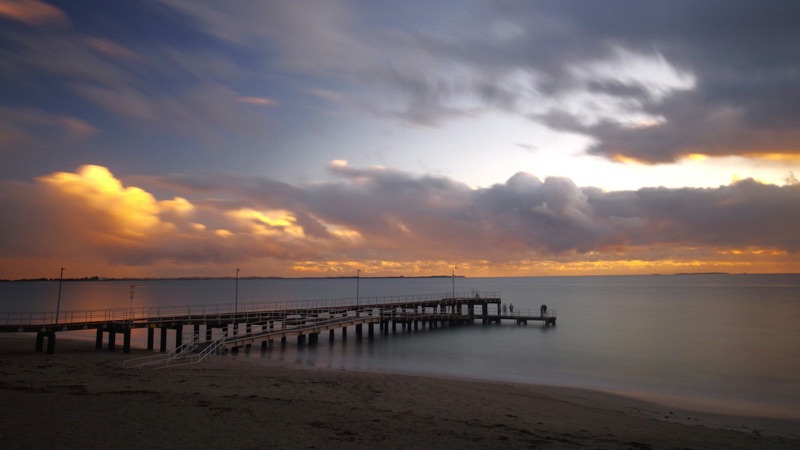 A wooden jetty reaching into calm water at dusk, a few figures at the far end.