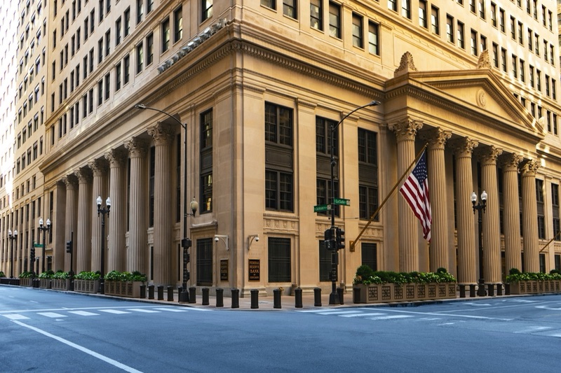 The columned stone corner entrance of the Federal Reserve Bank of Chicago building, with an American flag and trees along an empty street.