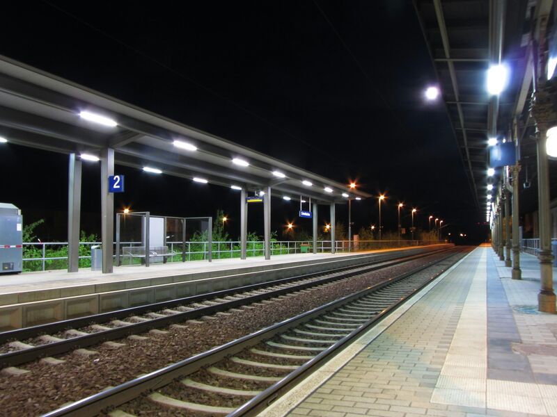 An empty railway platform at night, lit by overhead lamps, with tracks running into the dark.