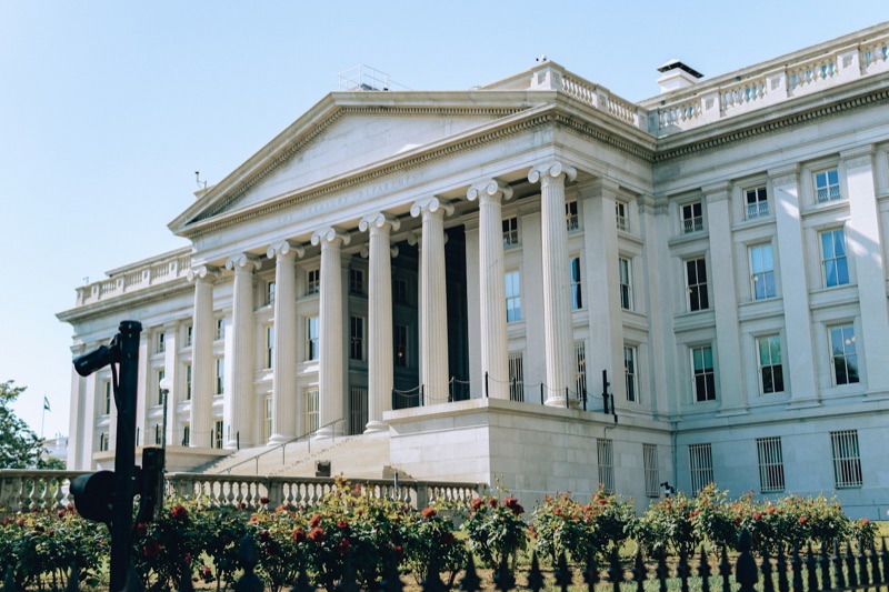 The columned portico of the Treasury Department building in Washington, with rose bushes and an iron fence in the foreground.