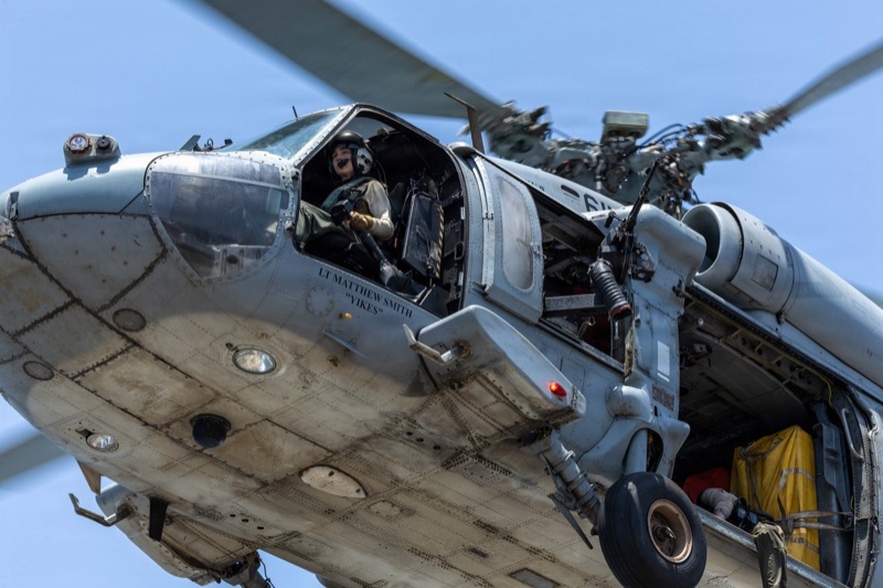 A grey U.S. Navy MH-60 Sea Hawk helicopter seen from below, banking against a blue sky, with a crew member seated in the open cockpit door beside a mounted machine gun.