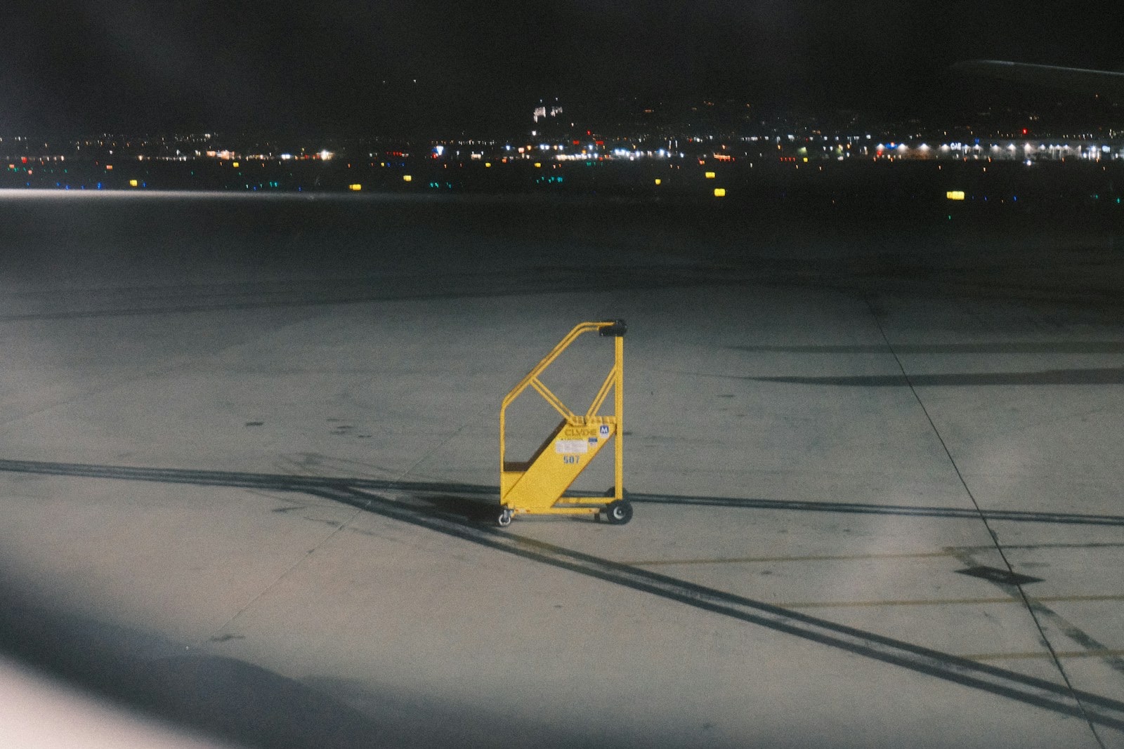 A yellow maintenance step-ladder alone on an airport apron at night, city lights on the horizon.