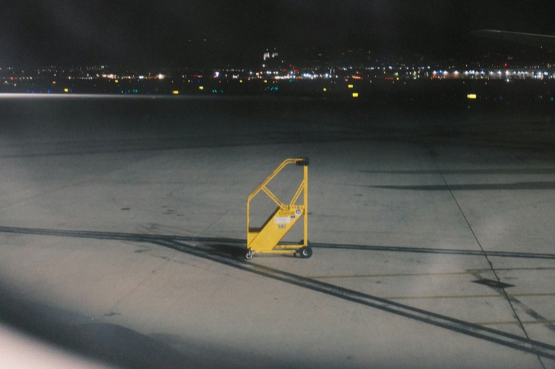 A yellow maintenance step-ladder alone on an airport apron at night, city lights on the horizon.