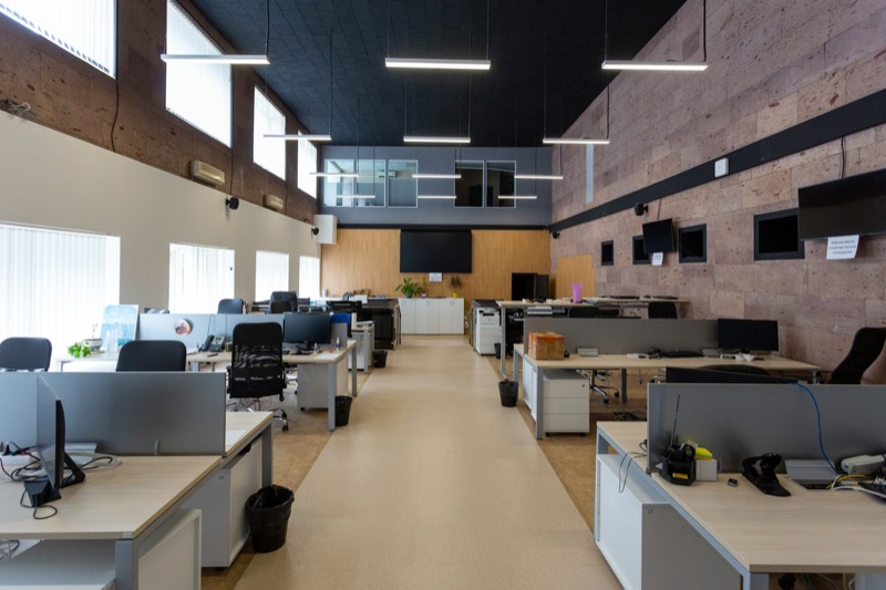 An empty open-plan office with two rows of pale desks running the length of the room, each with a monitor, keyboard and desk phone, under suspended linear lights and a dark ceiling.