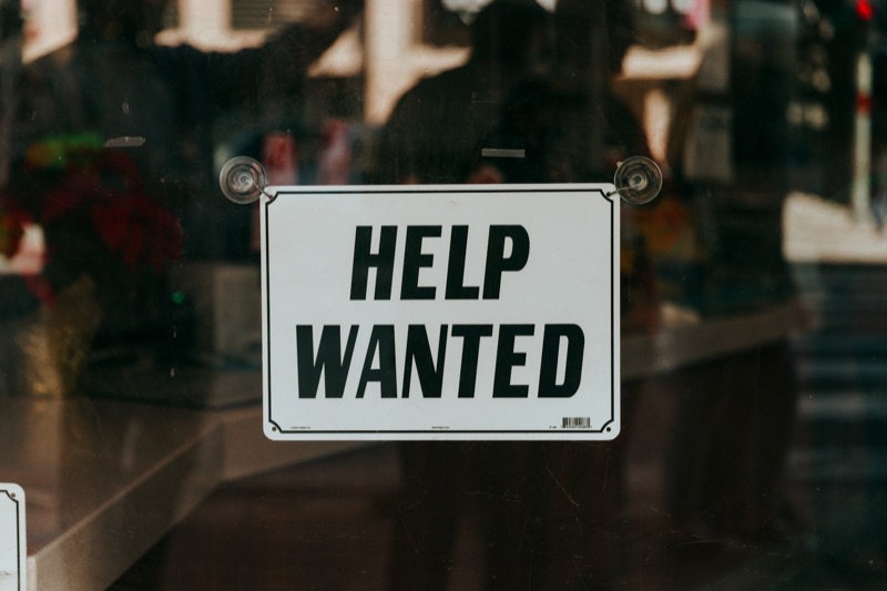 A white sign reading HELP WANTED in black capitals hangs by suction cups inside a shop's glass door, with blurred reflections of the street and people behind it.