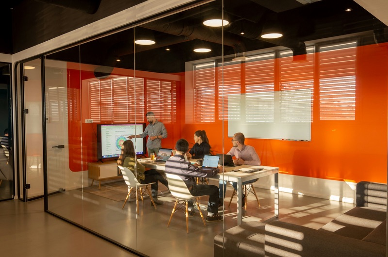 Five people seated and standing around a white table in a glass-walled meeting room with an orange feature wall, seen from the corridor outside through two panes of glass, laptops open and a chart on the screen at the head of the table.