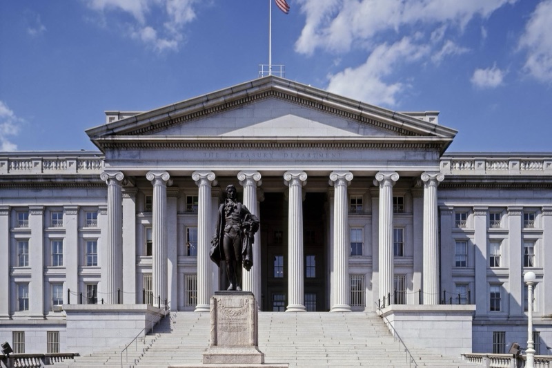 The columned south portico of the U.S. Treasury Department building in Washington, with a bronze statue of Alexander Hamilton on a stone pedestal in front and an American flag flying above the pediment under a blue sky.