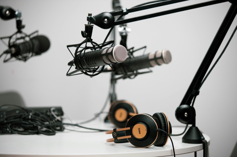 Three podcast microphones on boom arms over a white table, with two pairs of headphones and a coil of cables.