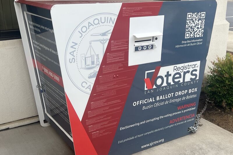 A red, white and navy San Joaquin County official ballot drop box chained outside a building, with bilingual English and Spanish signage and a QR code.