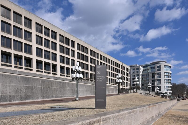 A dark sign reading "United States Department of Labor, 200 Constitution Avenue NW" stands on a lawn before the long, pale Frances Perkins Building under a blue sky with white clouds.