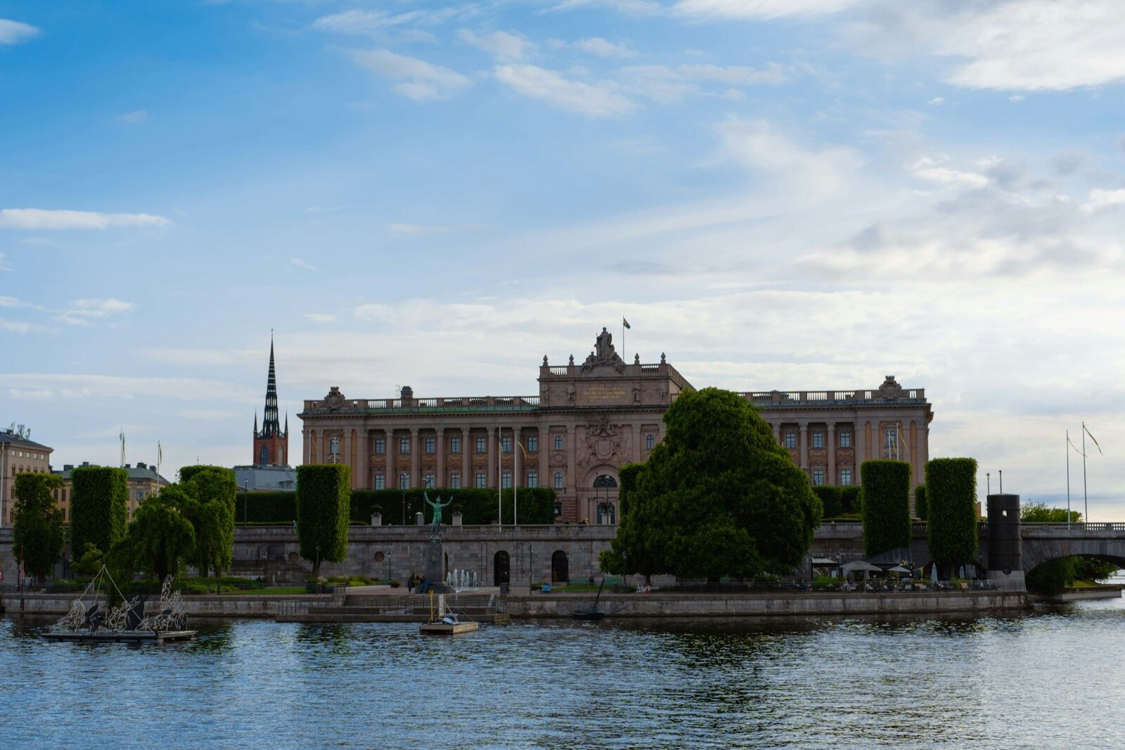 The Riksdag building in Stockholm seen across the water, with trees along the embankment and a church spire behind.