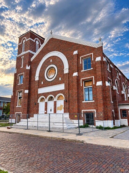 The red-brick facade of the former Euclid Methodist Church in St. Petersburg, Florida, with a white arched entrance, a round window and a square bell tower, behind a temporary chain-link fence on a brick street at sunset.