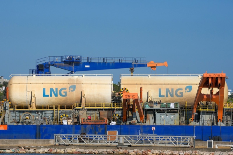 Two large cream-coloured cylindrical tanks marked LNG sit on a blue vessel moored at a quayside, with a blue and orange crane above them under a clear sky.