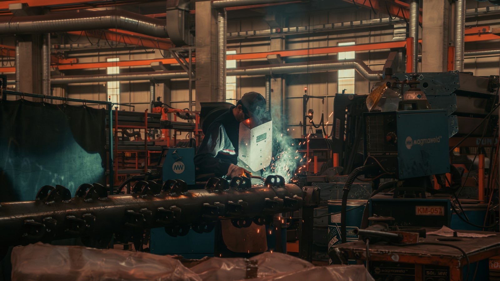 A welder in a helmet leans over a length of steel pipework in a metal fabrication shop, sparks and a bright blue arc flaring from the seam, with welding sets, gas bottles and overhead ducting filling the workshop behind him.