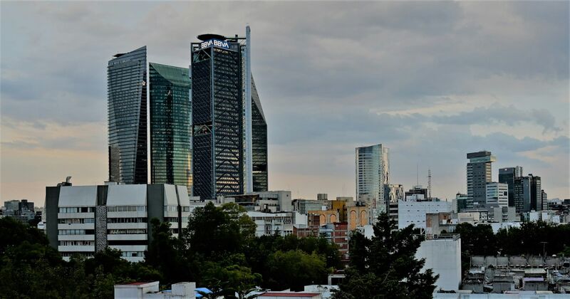 The Mexico City skyline seen over the rooftops of Colonia Condesa, with towers on Paseo de la Reforma under a cloudy sky.