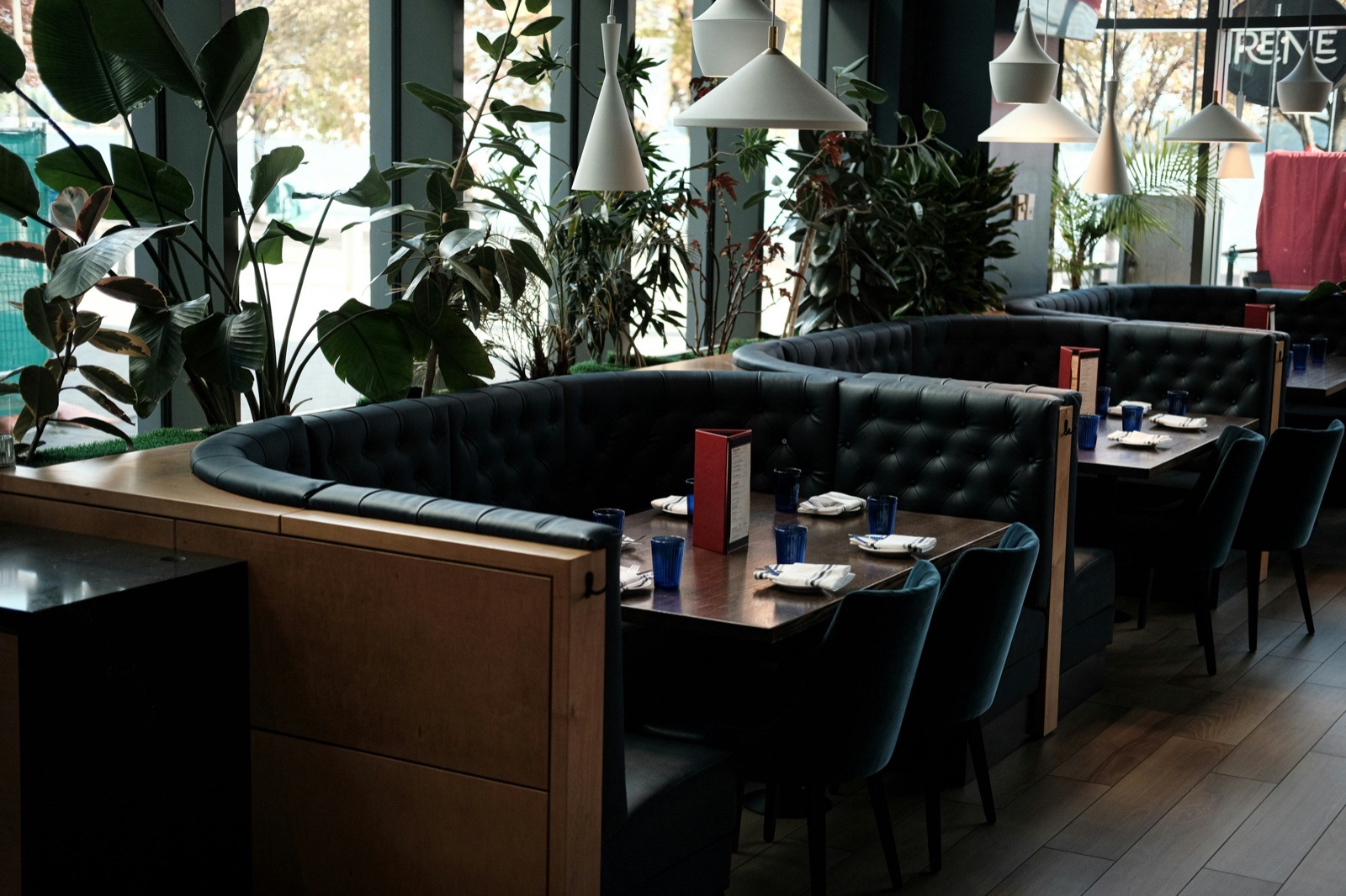 Empty dark leather booths and tables set for service in a restaurant dining room lined with large potted plants and pendant lamps.