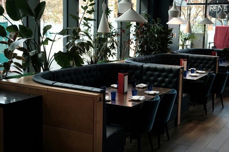 Empty dark leather booths and tables set for service in a restaurant dining room lined with large potted plants and pendant lamps.