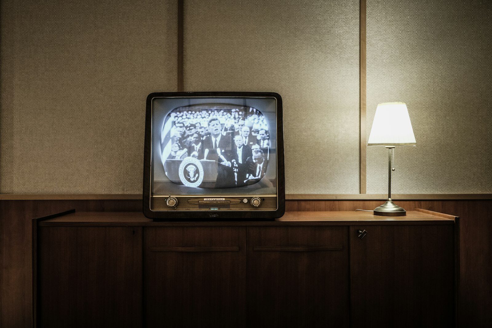 A vintage cathode-ray television on a wooden sideboard beside a lit table lamp, showing black-and-white archive footage of a press conference.