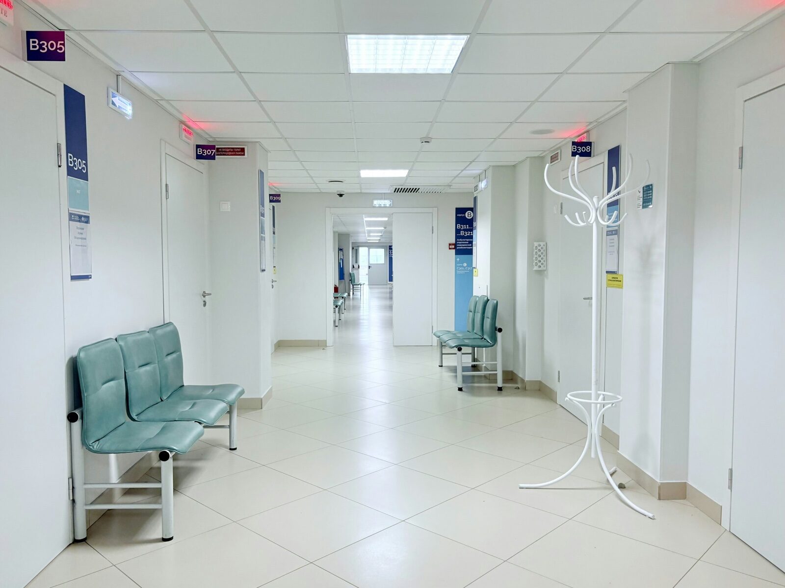 An empty, brightly lit clinic corridor with pale green benches and numbered doors.