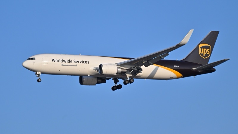 A UPS Boeing 767-300F freighter in brown and white livery, with 'Worldwide Services' on its fuselage and the UPS shield on its tail, flying with landing gear down against a clear blue sky.