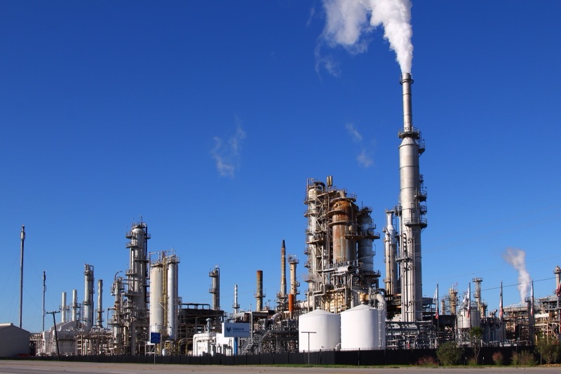 Distillation columns, storage tanks and a tall stack releasing white vapour at an oil refinery under a clear blue sky, with a Valero sign near the fence.