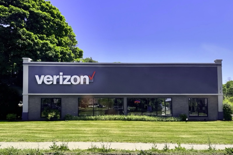 A single-storey brick store with a dark fascia bearing the white Verizon logo and red check mark, a row of glass windows with an Open sign, and a mown lawn in front, beside a large green tree.