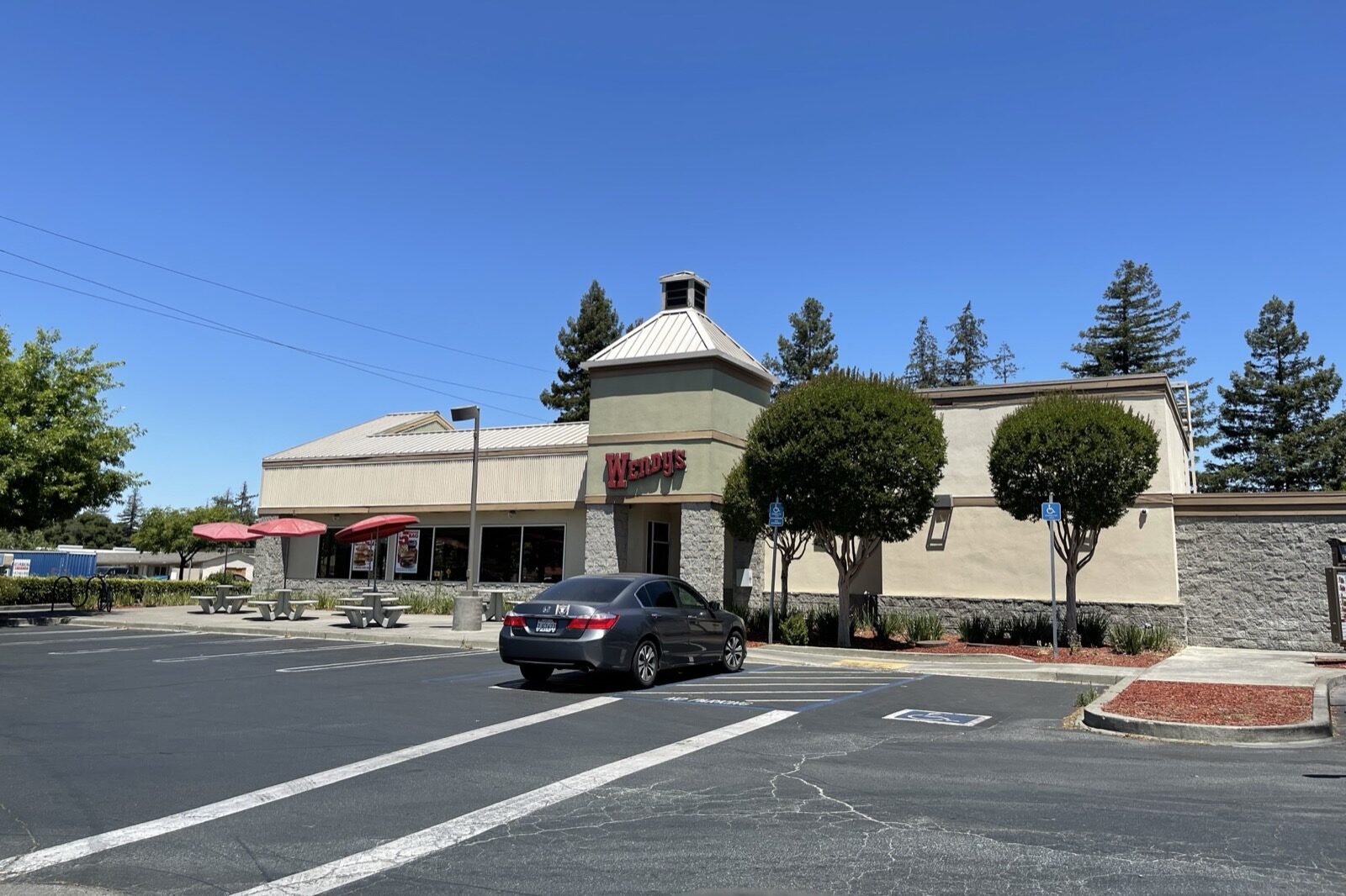 A Wendy's restaurant with a stone and stucco exterior, red patio umbrellas and a single grey car parked in front, under a clear blue sky.